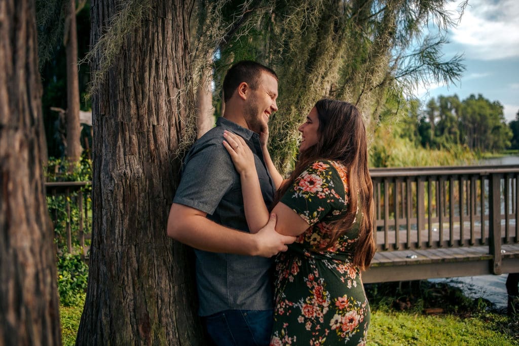 Engaged couple leaning against tree at Knotted Roots on the Lake